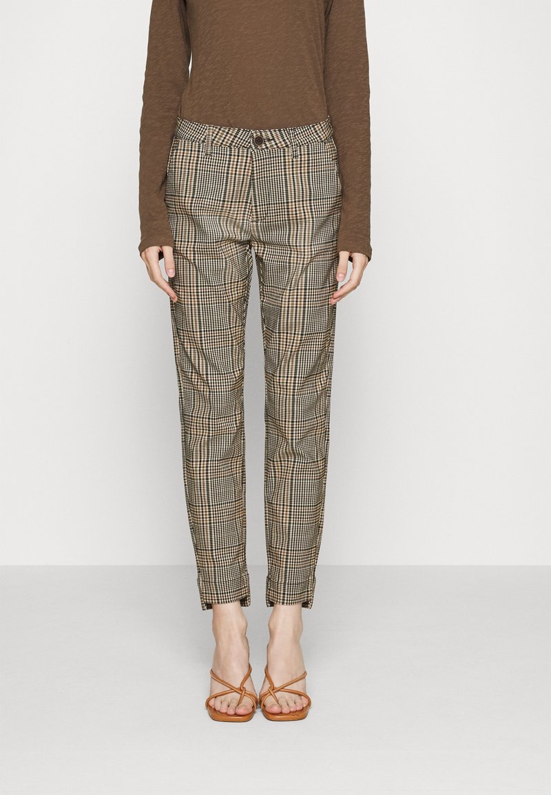 Pantalon ajusté beige à carreaux noirs, associé à un haut marron à manches longues et des sandales à talons fines couleur tan.