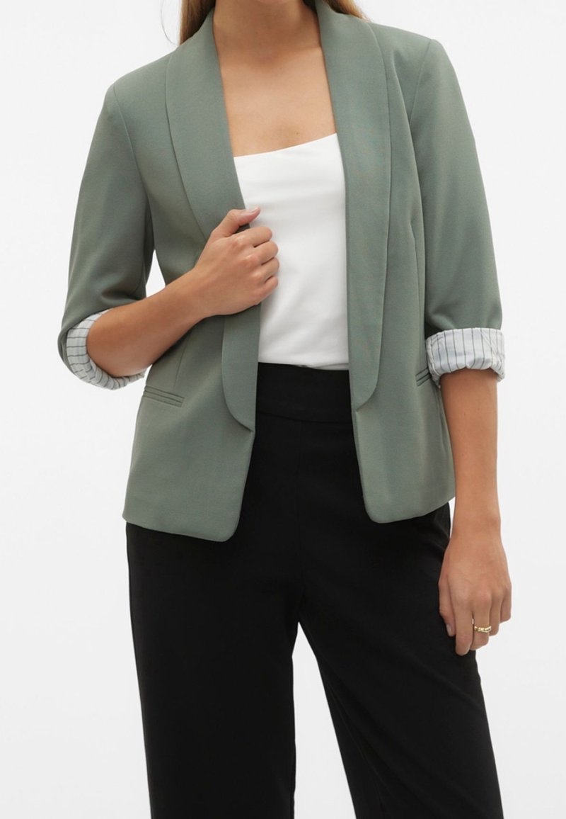 Femme portant un blazer vert sauge avec les manches retroussées, un top blanc sans manches et un pantalon noir, une main tenant le revers du blazer.