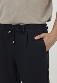 Męska dłoń w kieszeni czarnych spodni z gumką, w połączeniu z beżowym swetrem, podkreślająca fakturę materiału i swobodny styl.