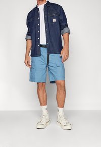 Man som bär en jeansskjorta över en vit t-shirt, ljusblå cargoshorts, vita högstövelskor och vita strumpor med "23" tryckt.