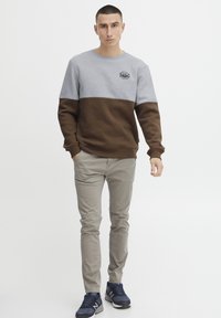 Grey and brown color-blocked sweatshirt with long sleeves, round neck, and textured fabric. Paired with light beige pants and dark sneakers.