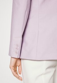 Liliowa marynarka z rękawami zapinanymi na trzy guziki, z dopasowanymi ramionami i czystym wykończeniem. Tkanina wydaje się gładka, z subtelną teksturą na jasnym tle.