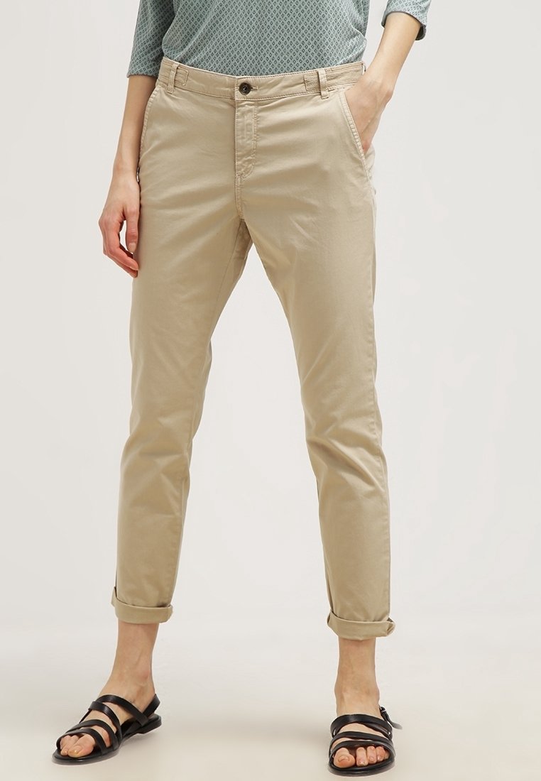 Personne portant un pantalon beige retroussé, un haut à manches longues à motifs et des sandales plates noires à brides, debout avec une main dans la poche.