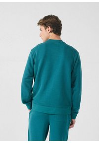 Teal crewneck sweatshirt van zacht stof, met geribbelde mouwen en zoom. De achterkant heeft een effen ontwerp zonder zichtbare logo's of patronen.