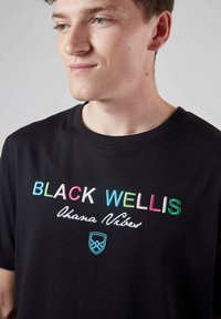T-shirt en coton noir avec du texte brodé multicolore : "BLACK WELLIS Ohana Vibes", avec un col rond simple et des manches courtes.