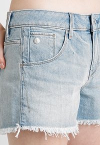 Pantalones cortos de mezclilla azul claro con dobladillo deshilachado, que incluyen un bolsillo frontal, un cierre de botón y detalles de costura. Textura suave.