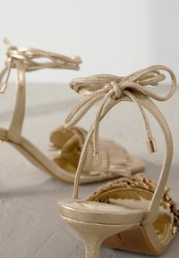 Gouden metallic hoge hakken sandaal met enkelbandjes, voorzien van franjes en een elegant ontwerp op een gestructureerd oppervlak.