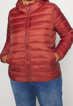 Personne avec les mains dans les poches, portant une doudoune rouge zippée et un jean bleu, cadrée au niveau du cou et des cuisses.