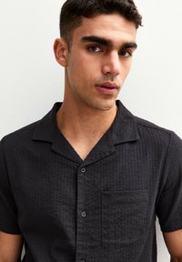Black short-sleeve shirt featuring a textured fabric, notched collar, front pocket, and black buttons.