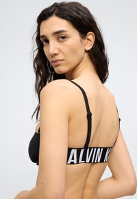 Γυναίκα με μακριά σκούρα μαλλιά που φοράει μαύρο σουτιέν Calvin Klein, κοιτάζοντας πάνω από τον ώμο της ενάντια σε ένα απλό φωτεινό φόντο.