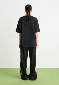 Black oversized T-shirt with short sleeves, paired with black track pants featuring yellow side stripes, viewed from the back.