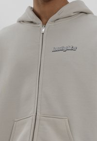 Ljusgrå hujacka med dragkedja, tillverkad av mjuk bomullsblandning. Har en kenguruficka och en "knowledge"-logga på bröstet i metalliskt silver.