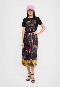 T-shirt grafico nero con un design colorato di tigre, indossato con una gonna maxi plissettata a fantasia floreale nei toni del nero e giallo, abbinato a sandali con laccetti.