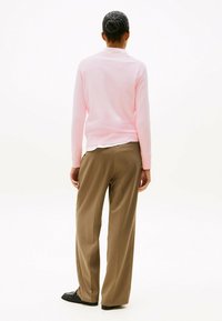 Blusenaus leichtem Rosa mit langen Ärmeln, lockere Passform, über einem weißen Hemd getragen. Olivgrüne weite Hose aus glattem Stoff, kombiniert mit schwarzen Schuhen.