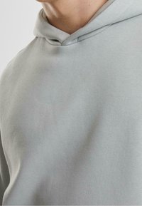 Närbild av en person som bär en ljusgrå huvtröja, med fokus på bröst- och nackområdet med huvan ner.