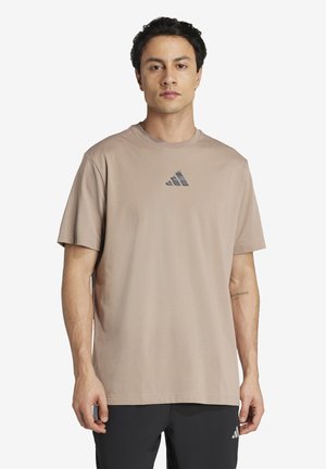 Homme debout, portant un t-shirt beige Adidas avec le logo centré et un pantalon noir avec un petit logo sur la cuisse, expression neutre, fond uni.