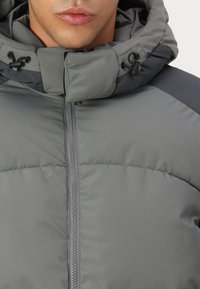 Grå pufferjakke med lynlås foran, høj krave og justerbar hætte. Har en blanding af matte og let shiny teksturer.