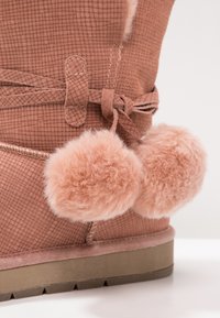 Růžové semišové kotníkové boty s texturovaným povrchem, zdobené dvěma huňatými bambulemi a kontrastní gumovou podrážkou. Součástí je nastavitelné vázání.