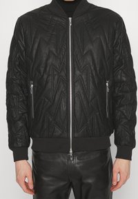 Veste bomber noire avec un motif en zigzag matelassé, fermeture éclair à l'avant, poches latérales et poignets côtelés. Fabriquée en tissu texturé avec une finition lisse.