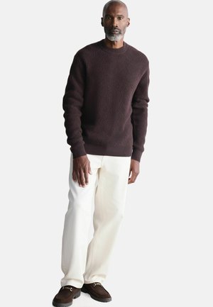Maglione a maglia marrone con texture e scollatura rotonda, abbinato a pantaloni bianchi a gamba dritta e scarpe in suede marrone scuro.