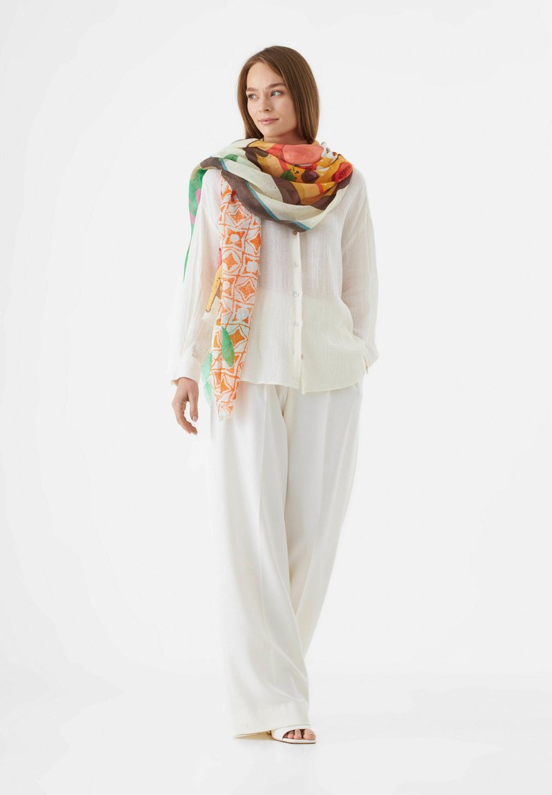 Chemise en lin blanche, ample, et pantalon large, associés à une écharpe colorée à motifs comprenant des oranges, des verts et des accents géométriques.