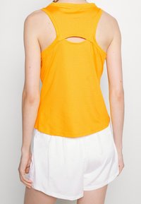 Top atlético sin mangas de color naranja con un acabado texturizado, que presenta un corte en forma de clave en la parte posterior y un dobladillo redondeado, combinado con pantalones cortos blancos.