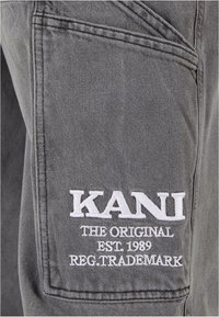 Tela gris con textura de mezclilla que presenta un logo bordado blanco que dice "KANI THE ORIGINAL EST. 1989 REG. TRADEMARK" en el bolsillo.