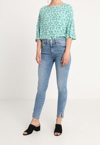 Leichte türkisfarbene Bluse mit floralem Muster, kombiniert mit enger sitzenden blauen Jeans mit ausgefranstem Saum und seitlichen Schlitzöffnungen sowie schwarzen Mules.