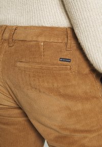 Bruine ribfluwelen broek met verticale ribbels, een achterzak met een logolabel en een gestructureerde afwerking. De tailleband is gestikt en voorzien van lussen voor een riem.