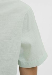 Camisa corta de manga verde claro con una tela texturizada. Presenta un corte relajado y un dobladillo limpio, mostrando una apariencia suave y transpirable.