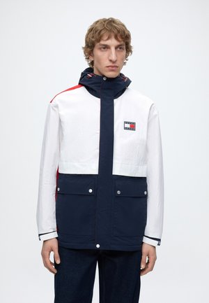 Mann mit blondem, lockigem Haar trägt eine farblich abgesetzte marineblaue, weiße und rote Kapuzenjacke mit Fronttaschen und Logo auf der Brust.