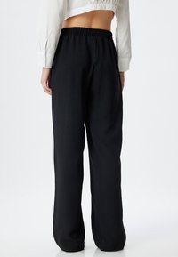 Zwarte, ruime sweatpants met een elastische tailleband, een gladde textuur en rechte pijpen, getoond van achteren tegen een effen achtergrond.