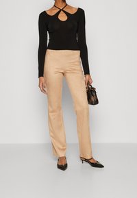 Top nero a maniche lunghe con apertura a chiave e pantaloni beige in finta pelle scamosciata a vita alta. Gli accessori includono una piccola borsa a tracolla con motivo e tacchi neri.