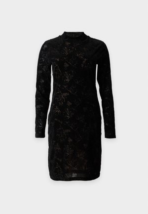 Femme aux cheveux bouclés, debout, portant une robe noire à manches longues longueur genoux et des sandales noires à talons hauts sur un fond gris uni.