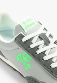 Graue Sneaker aus einer Mischung von Stoff und Wildleder, mit weißen Schnürsenkeln, grünem Totenkopf- und gekreuzten Knochen-Logo sowie einem markierten Zungenetikett.