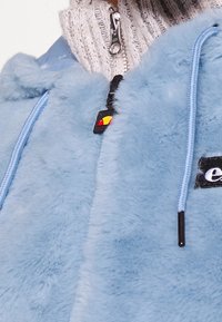 Jasnoniebieska kurtka ze sztucznego futra z wysokim kołnierzem, z czarnymi i żółtymi elementami na zamku błyskawicznym oraz niebieskimi troczkami.