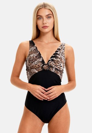 Kobieta o długich blond włosach, ubrana w jednoczęściowy kostium kąpielowy z dekoltem w serek, z panterkowym topem i czarnym dołem, z delikatnie splecionymi rękami na talii.