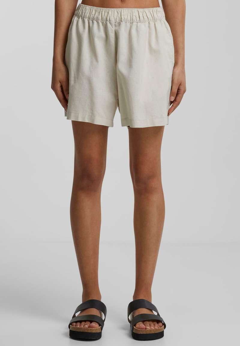 Shorts en lin beige avec une taille élastique, offrant une coupe ample et droite. Associés à des sandales à brides noires.