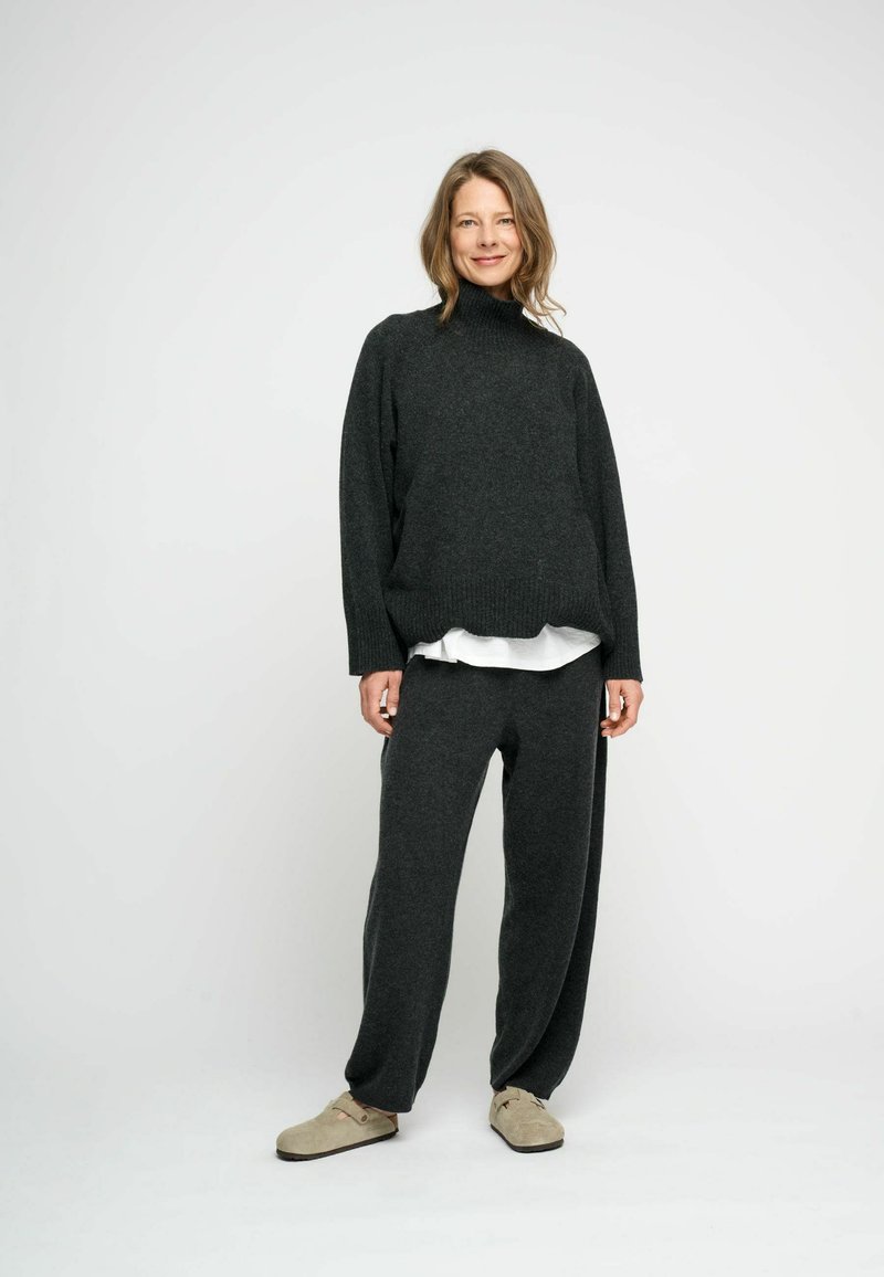Mörkgrå stickad tröja med hög krage, kombinerad med matchande löst sittande grå byxor och beige slip-on skor. Slät textur.