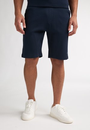 Pantalones cortos texturizados en azul marino con un diseño ajustado, de longitud por encima de la rodilla, combinados con zapatillas blancas. Presentan una cintura lisa y una marca mínima.