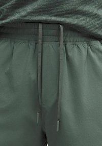Nahaufnahme von dunkelgrünen, elastischen Sweatpants mit passenden Kordeln und einem melierten grünen Shirt, das in die Hose gesteckt ist.