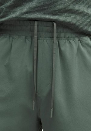 Gros plan sur un pantalon de survêtement à taille élastique vert foncé avec des cordons assortis et une chemise verte chinée rentrée à l'intérieur.