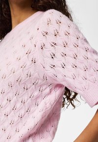 Gros plan d'une femme portant un pull en tricot rose clair avec un motif texturé ajouré en forme de feuilles et des manches courtes bouffantes.