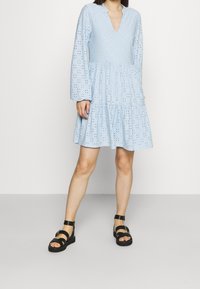 Abito azzurro chiaro, fino al ginocchio, con un corpetto strutturato e decorato e maniche lunghe. La gonna svasata presenta un design a strati. Abbinato a sandali neri.