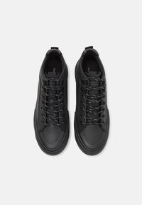 Ett par svarta låga sneakers med svarta och vita snören, släta tåkappor och dragflikar på vit bakgrund.