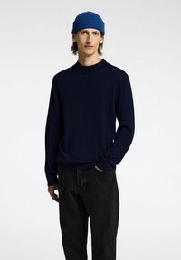 Maglione a maglia navy con collo rotondo, maniche lunghe e orli a costine, abbinato a pantaloni in denim neri e a un berretto blu brillante.