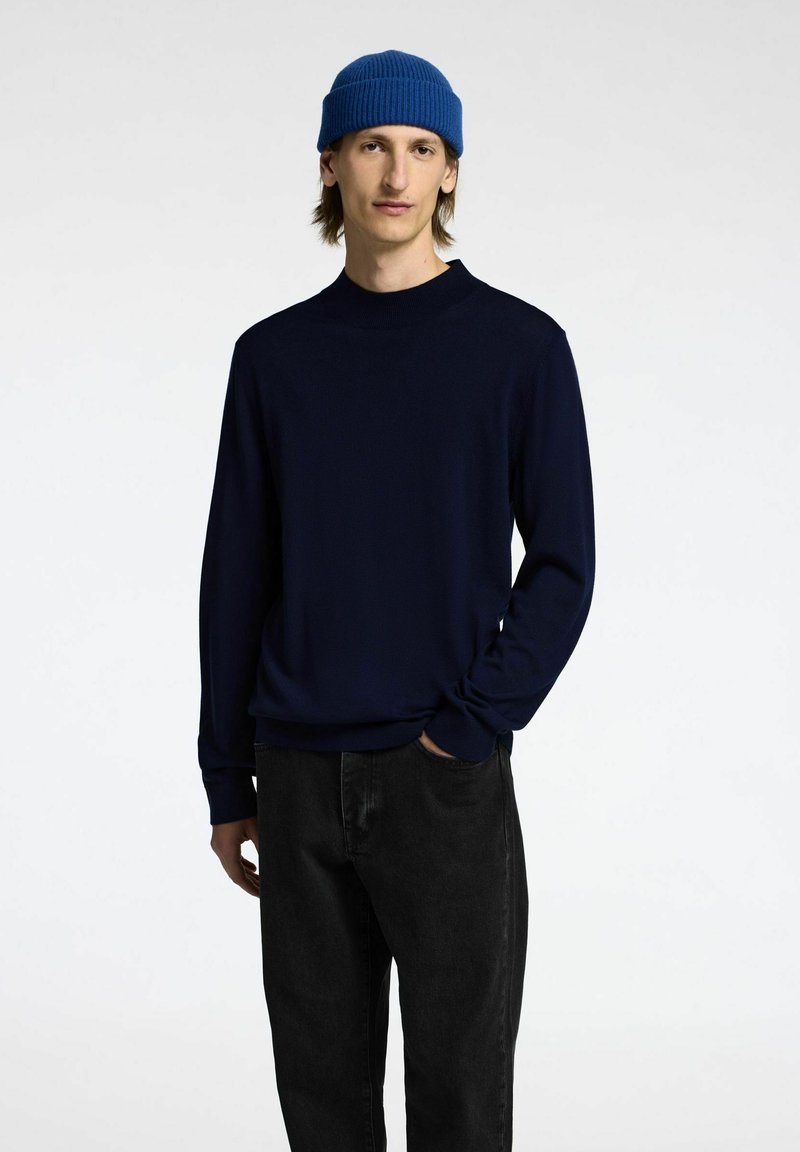Maglione a maglia navy con collo rotondo, maniche lunghe e orli a costine, abbinato a pantaloni in denim neri e a un berretto blu brillante.