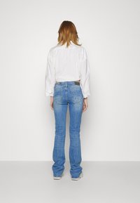 Jeans de ganga azul de cintura alta com um corte bootcut, apresentando um tom claro e um detalhe de bolso traseiro. Combinados com uma camisa branca.