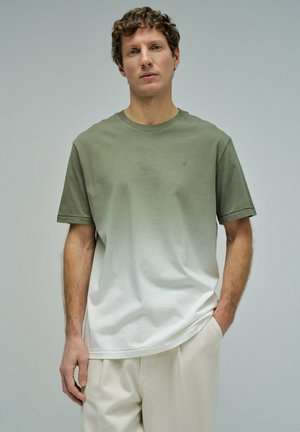 Hombre con el pelo corto y rizado que lleva una camiseta de degradado verde a blanco y un pantalón beige claro, de pie frente a un fondo liso y claro.