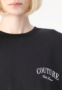 Juoda medvilninė marškinė su raukšlėta apykakle, priekyje su užrašu "COUTURE Belle Femme" atspausdintu kreivai balta spalva.